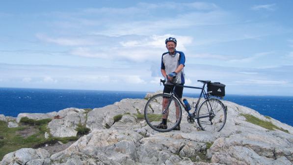 A man in blue and white cycling kit and helmet is standing on a rocky promontory holding a loaded touring bike. In the background is a very blue sea and blue sky