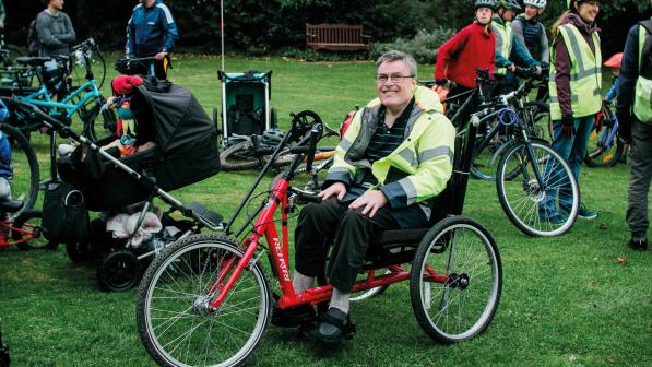 A man in a yellow hi-vis jacket, trousers and T-shirt is sitting on a red handcycle. He is smiling at the camera. There are several people in the background with lots of different types of cycle