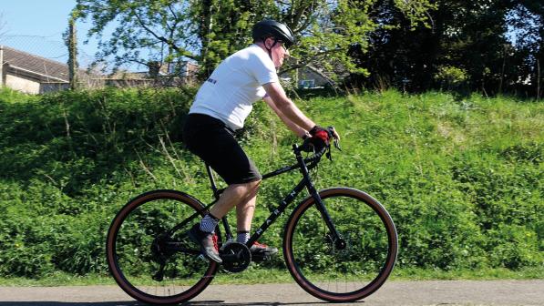 A man is riding a black touring bike along an off-road path. He is wearing a white T-shirt and black short, a cycling helmet and gloves