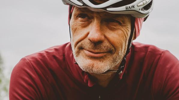 A man in a red long-sleeved cycling jersey and white cycling helmet with grey stubble is smiling and looking to the side of the camera