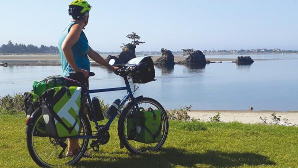 Woman in a cycling helmet and bluetop is standing holding a loaded touring bike looking out over a lagoon with large rocks in a line and a tree