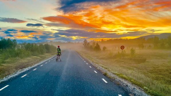 A person is standing astride a bike on a country road with trees and grass lining both sides. There is a spectacular sunset with the sky all orange, blue and yellow