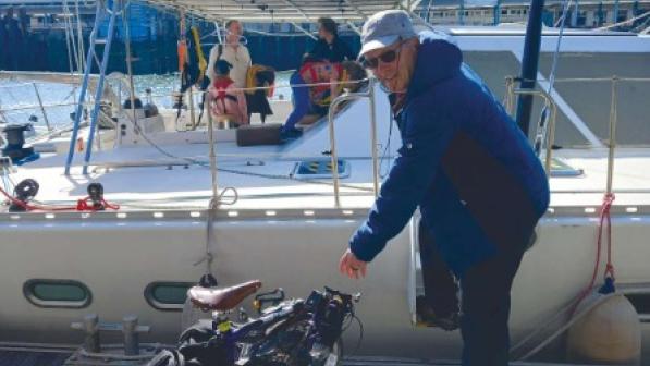 A man in blue coat, baseball cap and sunglasses is standing on a dock, bending down to pick up a folded Brompton bike. Behind him is a boat