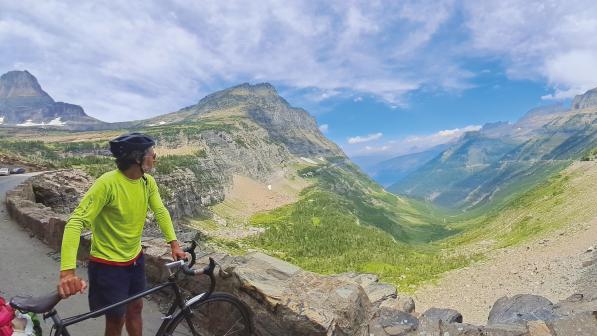 A man in bright yellow cycling jersey and blue shorts and helmet, holding a black touring bike, is looking out over a stunning mountain pass