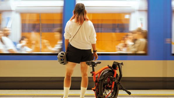 A woman in blue skirt and white top, with long hair and holding a cycle helmet, is standing on a train platform with a folded Brompton bike next to her as a train goes past