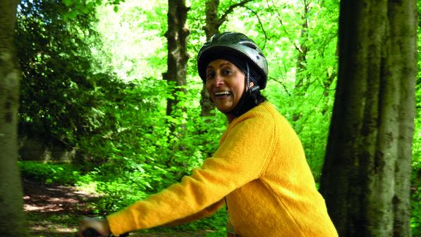 A woman in a yellow sweater and cycle helmet is holding onto the handlebar of a bike in a forest. She's smiling at the camera