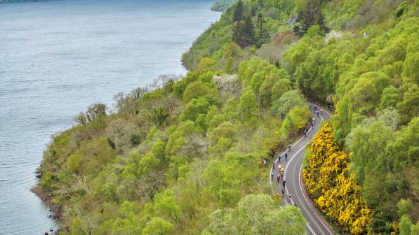 Taken from above, a line of cyclists are riding along a road that wends its way through a forest lining a loch with mountains in the background