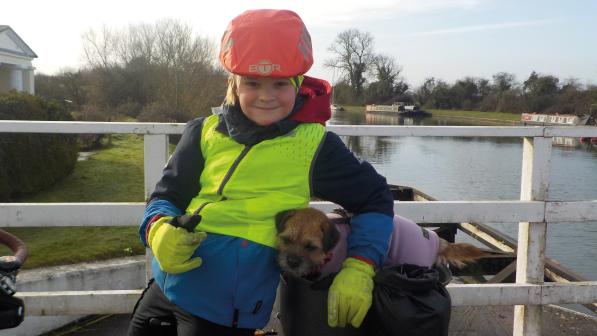 A small dog in a coat in a pannier bag on the back of a bike, with a child standing next to her with an arm around the dog. They're on a bridge and there's a river with boats on in the background