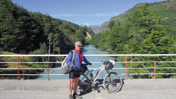 A man in cycling kit is standing with a loaded touring bike on a bridge over a river. Behind him are the river and pine-covered mountains underneath a blue sky