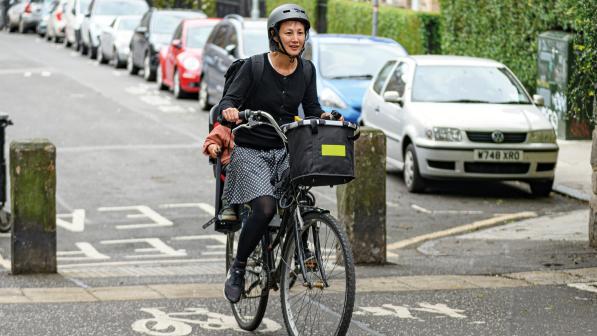 A woman is riding a Dutch-style bike with a basket on the front through a low-traffic neighbourhood. She has a child in a seat on the back. She is wearing a skirt and black sweater, tights and shoes and a cycle helmet