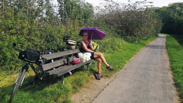 A woman in cycling kit is sitting on a bench next to a tarmac path. A bike is leaning against the back of the bench and she is holding a purple umbrella