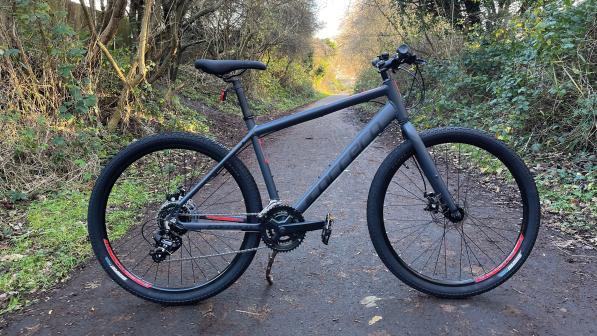 Carrera Subway 1, a dark grey hybrid bike with wide tyres and flat handlebar, propped up on a tree-lined off-road track