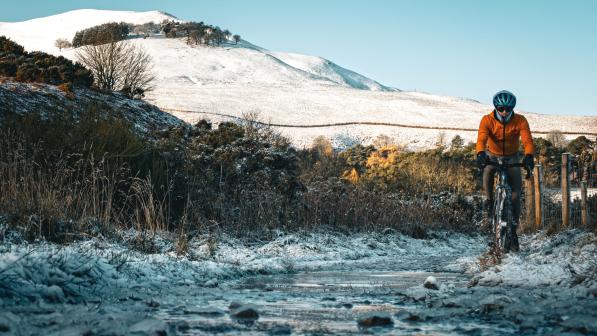 A person in an orange jacket is riding a gravel bike off road through snow and ice