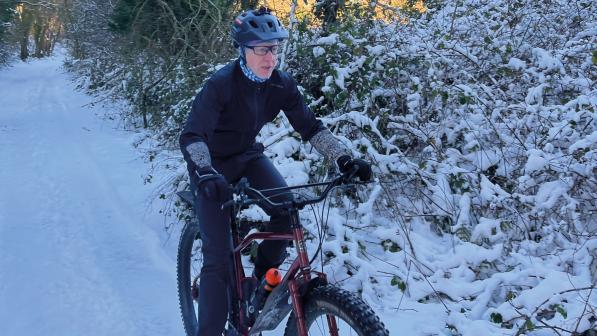 A man wearing warm winter cycling kit and a helmet is riding a mountain bike through snow in a wintery terrain