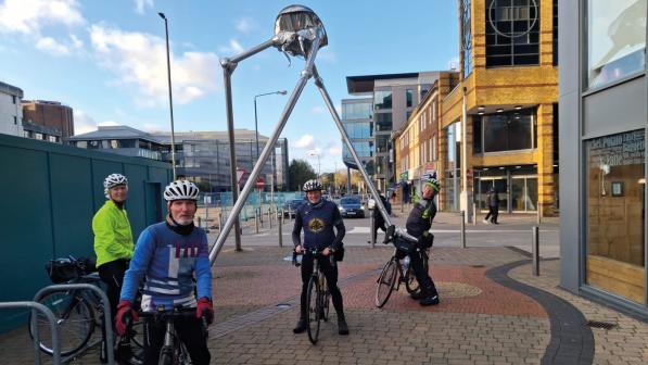 A group of four people in cycling kit and helmets are standing with their bikes in front of a metal statue of a Martian tripod from War of the Worlds