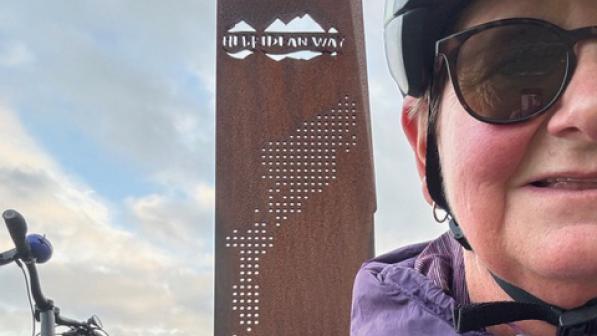 A woman in a purple waterproof jacket, sunglasses and cycle helmet is standing in front of a rusted metal sign with the words 'Hebridean Way' and a bike handlebar behind her