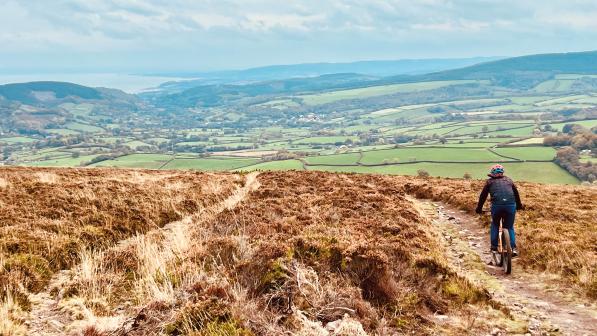 A person is riding a mountain bike on an off-road track at the top of a hill with a vista of hills and valleys and fields laid out in front