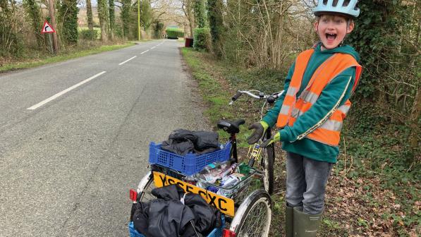 A boy in hi-vis jacket, jeans, wellies and green hoodie and a cycle helmet is indicating the treasures he's collected on his trike and smiling