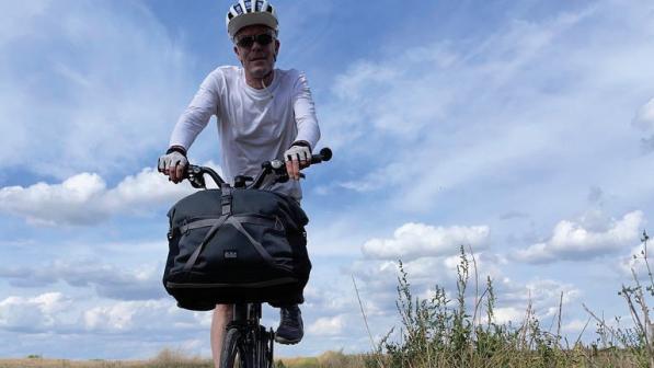 A man in a long-sleeved T-shirt, cycle helmet and sunglasses is riding a Brompton with a packed bag on the front on a gravel track