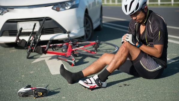 A man is sitting on the road with his legs out in front of him. One leg is bent and he's holding the knee, there is blood on his calf. He is wearing cycling kit. His bike is on the road behind him and behind that is a white car