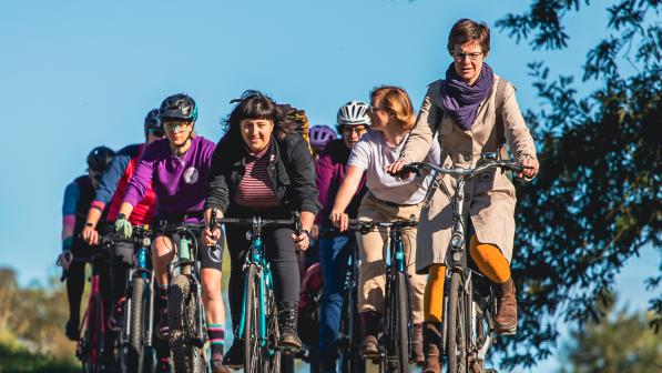 A big group of women is cycling on a tree-lined cycle path. They are wearing normal clothes and on a mix of bikes. They are smiling and chatting