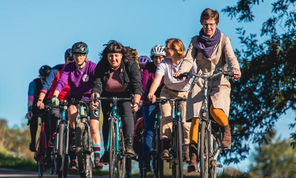 A big group of women is cycling on a tree-lined cycle path. They are wearing normal clothes and on a mix of bikes. They are smiling and chatting