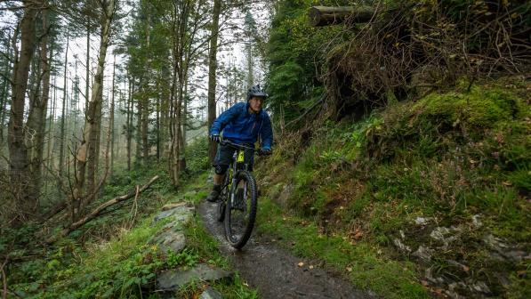 A man is cycling down a hill in a forest. He's on a mountain bike and is wearing waterproof cycling kit and a helmet