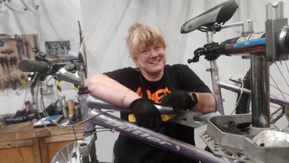 Cath Palgrave is working on a light metallic mauve Claud Butler mountain bike in a mechanics’ workshop. The bike is clamped to a work stand. Cath is wearing a black T-shirt and black gloves. She has blonde hair and hoop earrings. She is smiling at the camera.