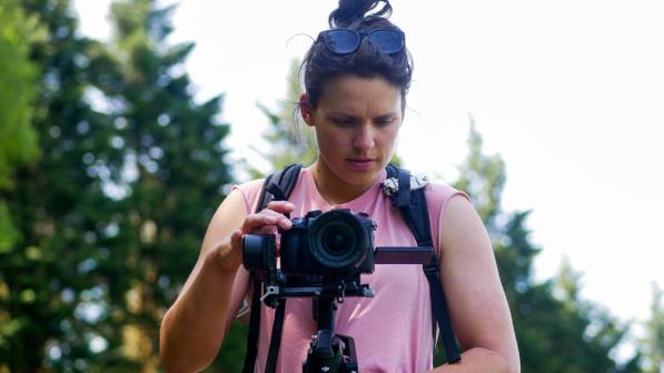 Filmmaker Catherine Dunn peering into her camera in a woodland. She is wearing a pink sleeveless t-shirt and a rucksack.