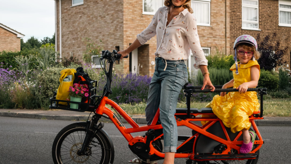 Caz Conneller is sitting astride an orange longtail cargo bike. She is wearing three-quarter-length blue jeans and a shirt. She has a helmet on. A little girl is sitting on the back of the bike. She is wearing a yellow party dress and patterned helmet. Two bags and a pot plant are in the basket at the front.