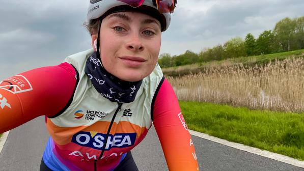 Charlotte Broughton has taken a selfie as she rides along a road. She is wearing race kit: a UCI Women’s Continental Team jersey and red arm warmers, a blue buff and white helmet. She has cycling sunglasses tucked into her helmet.