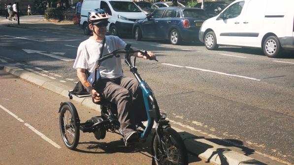A man in a white T-shirt and trousers, wearing a cycle helmet and sunglasses, is riding a trike on protected cycle lanes next to a road