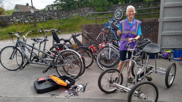 Christine Fisher is standing surrounded by lots of bikes, including tandems, trikes and quads. There is a big toolbox open on the ground and she is holding up a tool with her other hand on a silver quad bike.