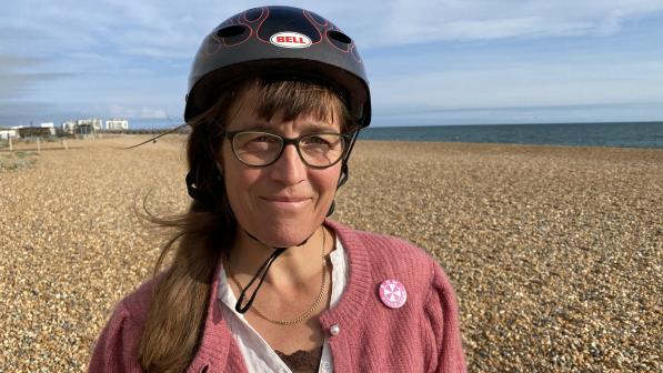Cicely Lloyd is standing on a pebble beach with the sea in the background. She is wearing a black Bell helmet, a pink cardigan and white shirt. She has a BMW Cycle Club badge on her cardigan. She has long brown hair and glasses. She is smiling at the camera.