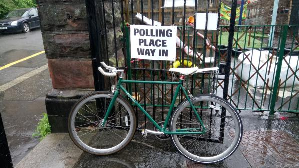 A green fixed-wheel bike is leaning against a black gate with a sign on it that reads 'Polling place, way in'