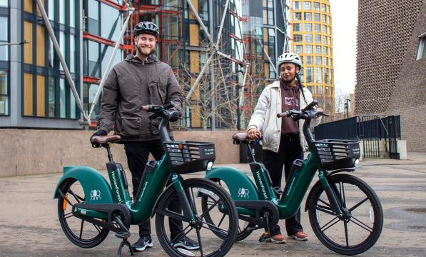 Two people are standing holding Human Forest e-bikes, green bikes with a black basket on the front and with the Human Forest logo. They are in an urban environment and wearing normal clothes and bike helmets.