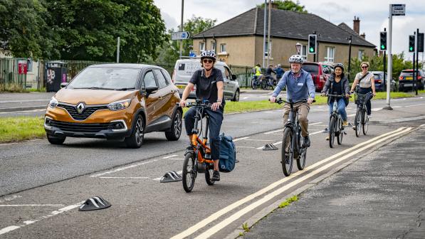 A group of people on different bikes is riding on a cycle lane separated from the road by armadillos. They're all wearing normal clothes and there's an orange car on the road behind them