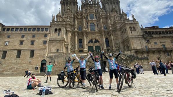 Four women in matching cycling kit are standing with loaded touring bikes in front of a church with their arms up in the air in a gesture of celebration