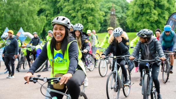 A group of cyclists of diverse backgrounds take part in a community cycling event in a park. A woman in the foreground, wearing a helmet and a bright yellow vest with "No Limits No Health" printed on it, smiles while riding her bike. Other participants, also wearing helmets and warm clothing, follow closely behind. The atmosphere is lively, with people engaging in cycling activities in an open, green space.