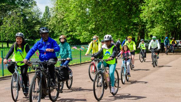 A group of adults ride bikes together through a tree-lined park in Wolverhampton during a Big Bike Revival event. Many wear high-visibility vests and helmets, smiling as they cycle in the sunshine.