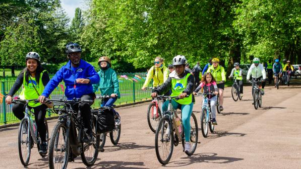 A group of adults ride bikes together through a tree-lined park in Wolverhampton during a Big Bike Revival event. Many wear high-visibility vests and helmets, smiling as they cycle in the sunshine.