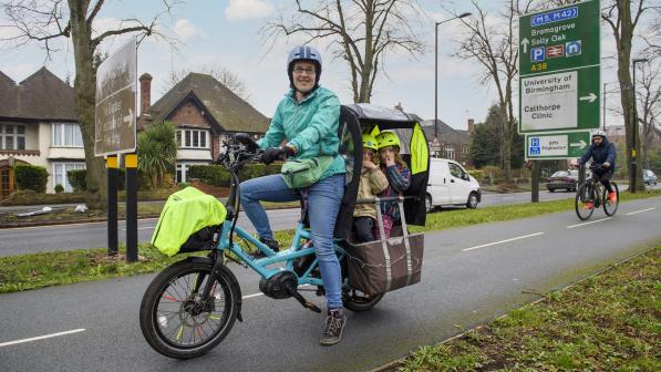 A woman is sitting on a turquoise e-cargo bike. There are two children on the back. She's wearing jeans and a turquoise jacket and cycle helmet. It's winter and there are no leaves on the trees
