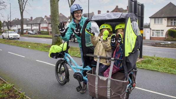 A woman is standing next to a pale blue cargo bike on a paved cycle path that is separated from the road. There are two young children in the cargo box. They are all holding up their hands to the camera to show their gloves which have smiley faces on them