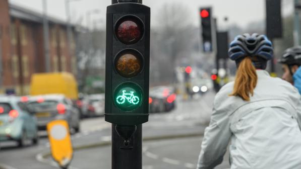 A close-up of a bike traffic light with the green bike lit up. In the background is a person on a bike, wearing a cycle helmet and waterproof jacket.