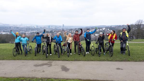 A row of people stand with bikes on a hill with a view over Sheffield in the background. They are smiling and waving at the camera. In the centre is Louise Haigh, Secretary of State for Transport
