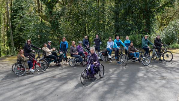 A group of adults in a park on a variety of non-standard cycles, tandems, e-assist cycles, handcycles and tricycles