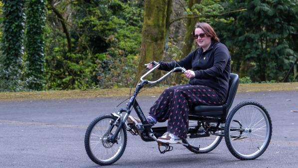 A woman is riding a tricycle in a park. She is wearing normal clothing and smiling