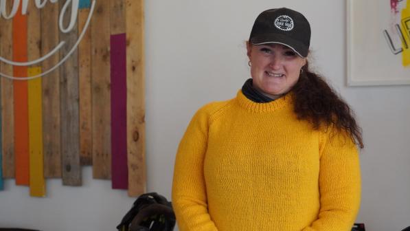 A woman wearing a bright yellow jumper and a black "Bike Hub" cap smiles at the camera. She stands indoors, in front of a wooden wall feature with colorful vertical planks and the word “unity” partially visible. Two bike helmets are visible in the background