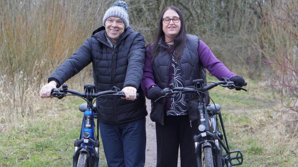 Two people are holding electric bikes and standing together on a muddy path with trees and bushes in the background. They're wearing warm clothing and are both smiling