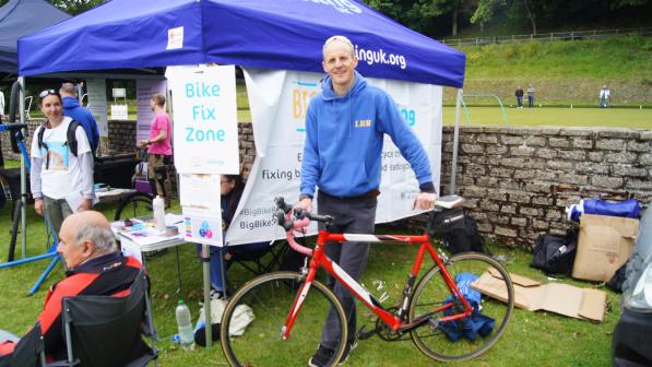 A smiling man in a blue hoodie stands holding a red road bike in front of a Big Bike Revival tent marked "Bike Fix Zone", surrounded by tools, boxes and event attendees on a grassy field.
